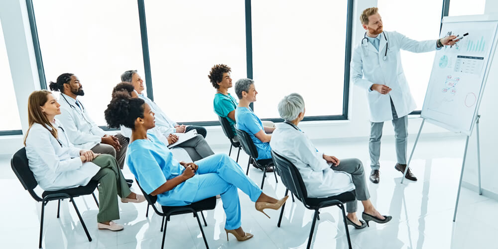 A nurse leader standing at the front of a board, addressing a group of staff members.