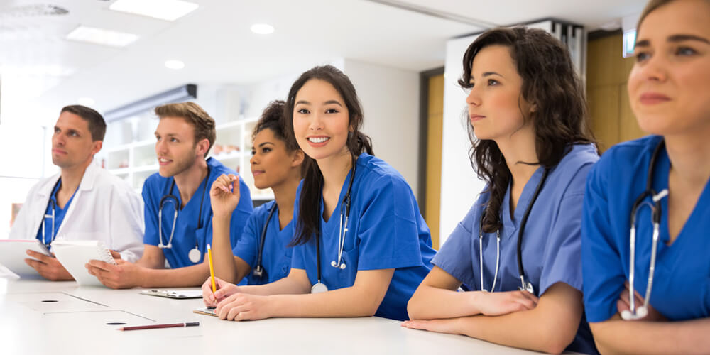 A nurse educator stands at the front of a classroom, in front of a group of nurses.
