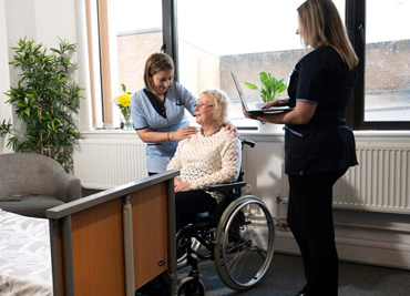 A senior citizen in a wheelchair is assisted by two healthcare workers